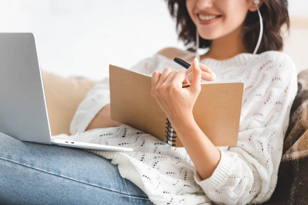 Обрізаний вид усміхненої дівчини, що пише в блокноті і вивчає онлайн з ноутбуком — стокове фото