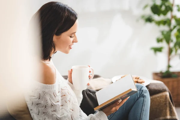 Glückliche Frau liest Buch auf Sofa mit Kaffee und Keksen — Stockfoto