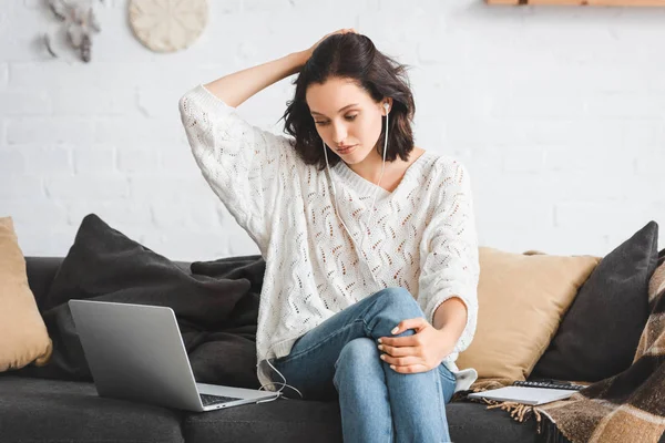 Attractive girl thinking and studying online with notepad, earphones and laptop on sofa — Stock Photo