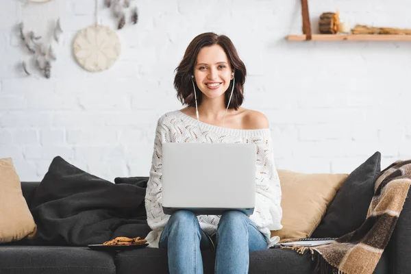 Smiling girl using earphones and laptop on sofa with cookies — Stock Photo