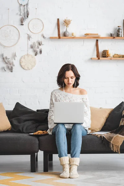 Konzentrierte Frau mit Kopfhörer und Laptop auf Sofa mit Keksen — Stockfoto
