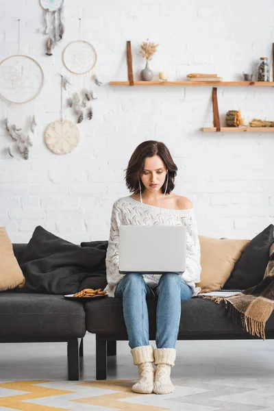 Femme concentrée utilisant écouteurs et ordinateur portable sur canapé avec des cookies — Photo de stock