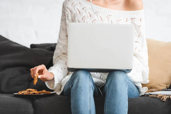 Vue recadrée de femme manger des biscuits avec ordinateur portable sur le canapé à la maison — Photo de stock