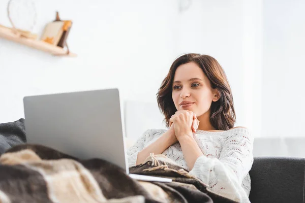 Mujer joven feliz en manta usando el ordenador portátil en el sofá en la acogedora sala de estar — Stock Photo