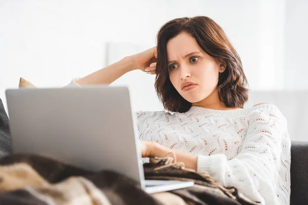 Beautiful angry girl in blanket using laptop on sofa in cozy living room — Stock Photo