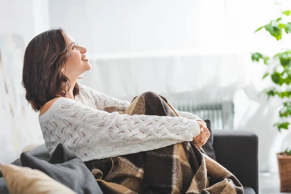 Счастливая брюнетка в одеяле, сидящая на диване в уютной гостиной — стоковое фото