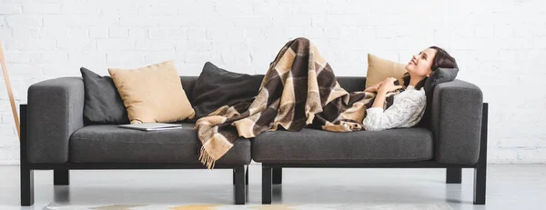 Menina morena feliz em cobertor deitado no sofá com laptop — Fotografia de Stock