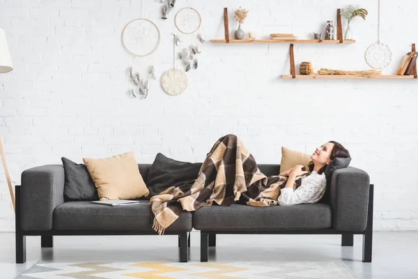 Menina bonita feliz em cobertor deitado no sofá com laptop na acolhedora sala de estar — Fotografia de Stock