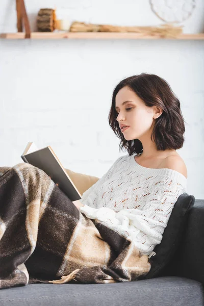 Beautiful girl in blanket reading book on sofa at home — Stock Photo