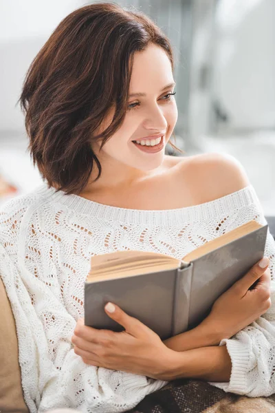 Hermosa mujer sonriente leyendo libro en el sofá en casa - foto de stock