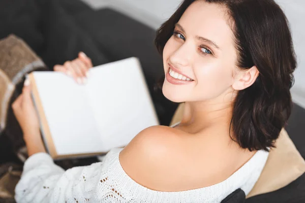 Красивая улыбающаяся девушка читает книгу на диване дома — стоковое фото