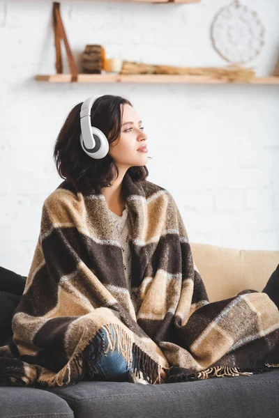 Beautiful girl thinking and listening music with headphones while sitting in blanket — Stock Photo