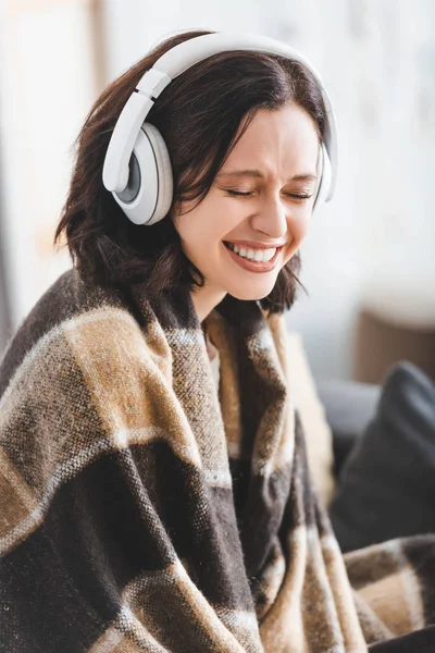 Hermosa mujer sonriente con los ojos cerrados en manta escuchando música con auriculares - foto de stock