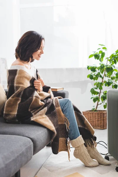Beautiful happy girl with blanket warming up with heater in cold room — Stock Photo