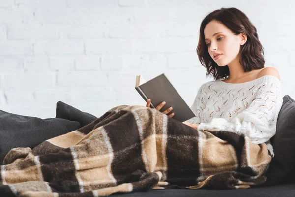 Belle jeune femme dans couverture livre de lecture sur canapé à la maison — Photo de stock