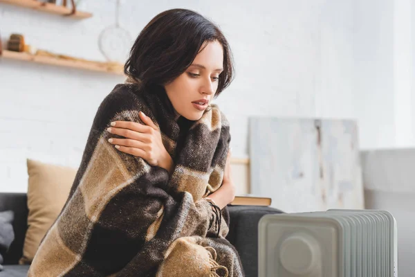 Beautiful girl with blanket warming up with heater in cold room — Stock Photo