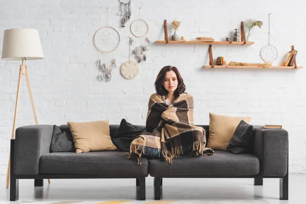 Attractive cold girl warming up with blanket on sofa in living room with dream catchers — Stock Photo