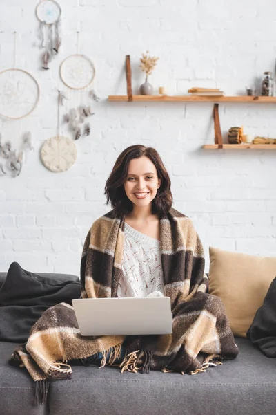 Menina bonita feliz em cobertor usando laptop no sofá na sala de estar com coletores de sonhos — Fotografia de Stock