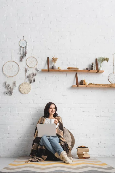 Cheerful brunette girl with cookies and coffee using laptop in living room with dream catchers — Stock Photo