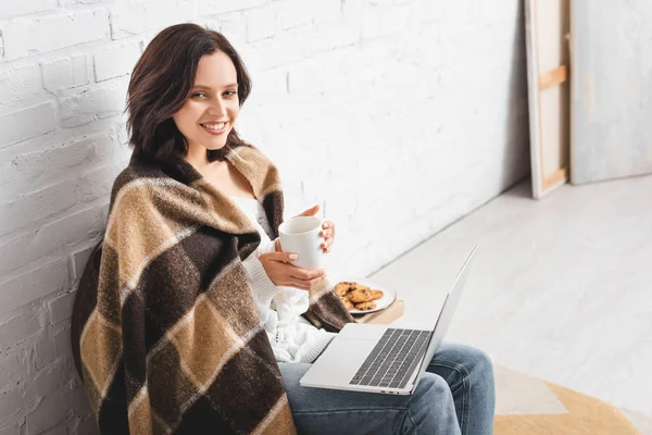Красивая улыбающаяся девушка в одеяле с печеньем и кофе с помощью ноутбука — стоковое фото