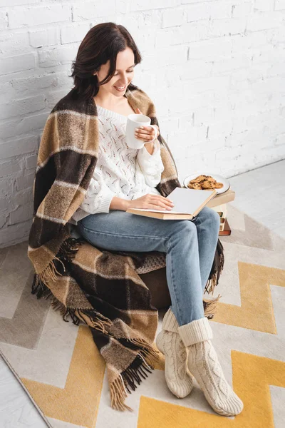 Happy girl in blanket reading book in cozy living room with coffee and cookies — Stock Photo