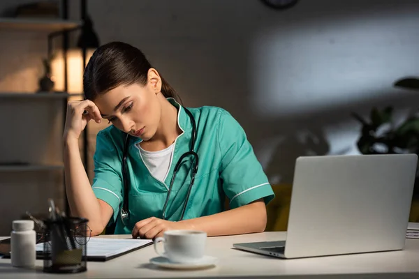 Attraktive und müde Krankenschwester in Uniform am Tisch sitzend und während der Nachtschicht auf Klemmbrett schauend — Stockfoto
