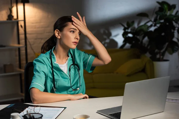 Enfermera atractiva y cansada en uniforme sentada en la mesa y mirando hacia otro lado durante el turno de noche — Stock Photo