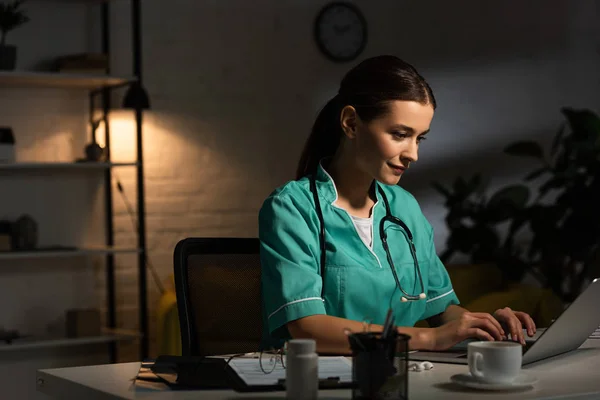 Enfermeira atraente em uniforme sentado à mesa e usando laptop durante o turno da noite — Fotografia de Stock