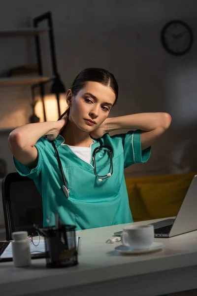 Attractive nurse in uniform sitting at table and having neck pain during night shift — Stock Photo