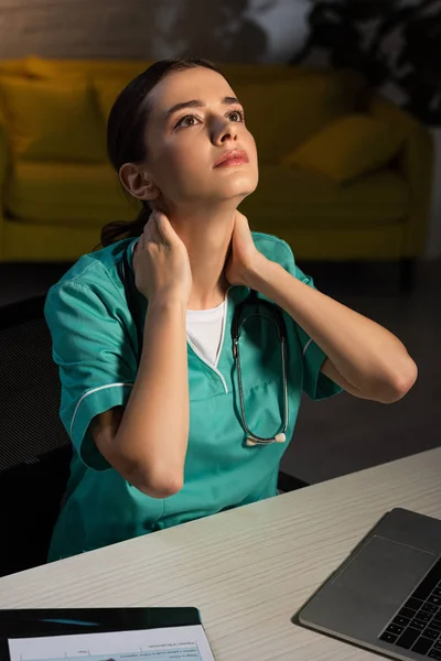 Infirmière attrayante en uniforme assis à table et ayant mal au cou pendant le quart de nuit — Photo de stock