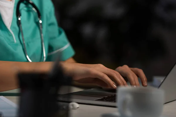 Vista cortada de enfermeira em uniforme sentado à mesa e usando laptop durante o turno da noite — Fotografia de Stock