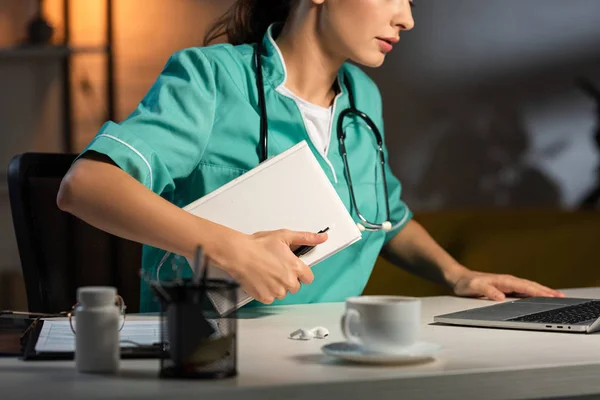 Vista recortada de la enfermera en uniforme sentada en la mesa y sosteniendo el cuaderno durante el turno de noche - foto de stock