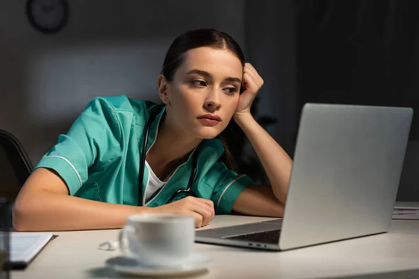 Attraktive Krankenschwester in Uniform, die während der Nachtschicht am Tisch sitzt und auf den Laptop schaut — Stockfoto