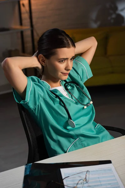 Attraktive Krankenschwester in Uniform sitzt während der Nachtschicht mit verschränkten Armen am Tisch — Stockfoto
