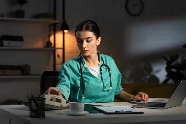 Attraktive Krankenschwester in Uniform sitzt am Tisch und nimmt Hörer während der Nachtschicht — Stockfoto