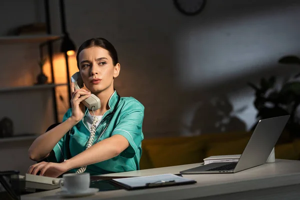 Привлекательная медсестра в форме сидит за столом и разговаривает по телефону в ночную смену — стоковое фото