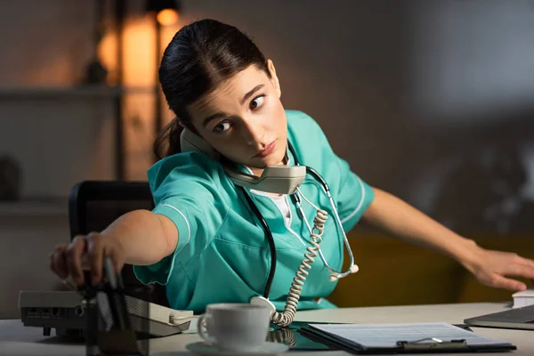 Attraktive Krankenschwester in Uniform sitzt am Tisch und telefoniert während der Nachtschicht — Stockfoto