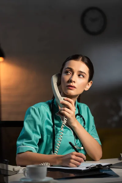 Enfermera atractiva en uniforme sentada en la mesa y hablando por teléfono durante el turno de noche - foto de stock