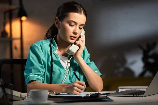 Attractive nurse in uniform talking on telephone and writing in clipboard during night shift — Stock Photo