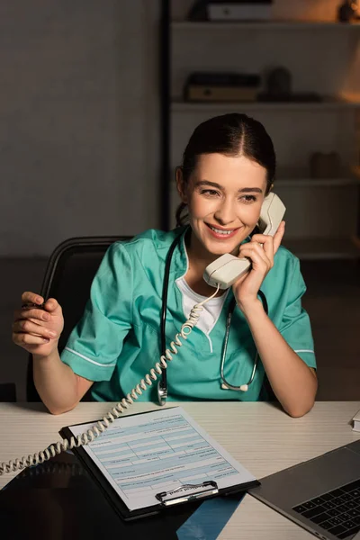 Infirmière souriante en uniforme assise à table et parlant au téléphone pendant le quart de nuit — Photo de stock