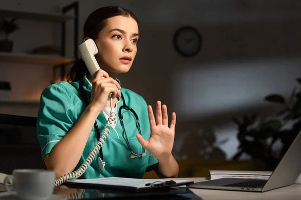 Шокована медсестра в уніформі сидить за столом і розмовляє по телефону під час нічної зміни — стокове фото