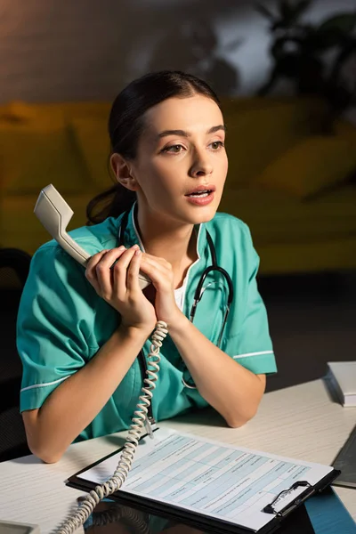 Enfermera atractiva en uniforme sentada en la mesa y sosteniendo el teléfono durante el turno de noche - foto de stock