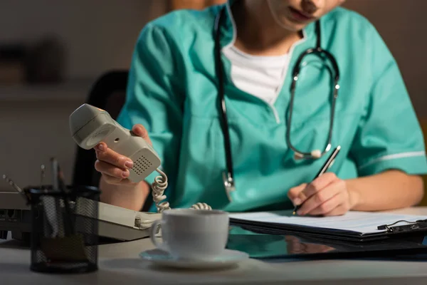 Ausgeschnittene Ansicht einer Krankenschwester in Uniform, die während der Nachtschicht schreibt und Hörer hält — Stockfoto