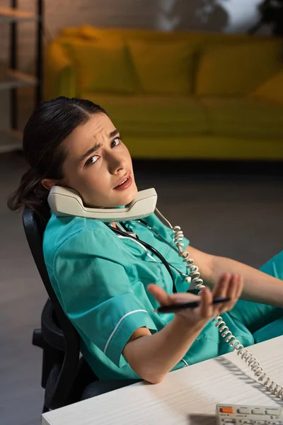 Infirmière attrayante en uniforme assise à table et parlant au téléphone pendant le quart de nuit — Photo de stock