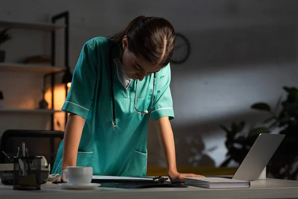 Attractive nurse in uniform with stethoscope looking down during night shift — Stock Photo