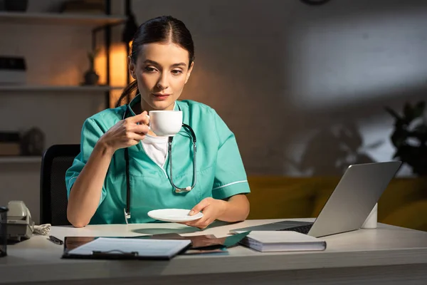 Attractive nurse in uniform sitting at table and drinking coffee during night shift — Stock Photo