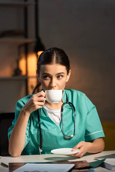 Attraktive Krankenschwester in Uniform sitzt am Tisch und trinkt während der Nachtschicht Kaffee — Stockfoto