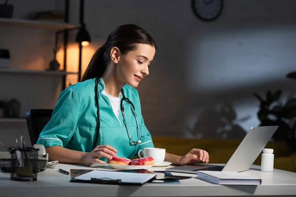 Attraktive und lächelnde Krankenschwester in Uniform, die während der Nachtschicht am Tisch sitzt und auf den Laptop schaut — Stockfoto
