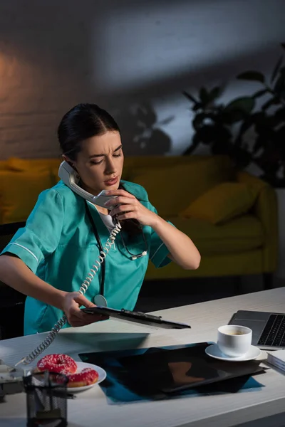 Nurse in uniform talking on telephone and holding clipboard during night shift — Stock Photo