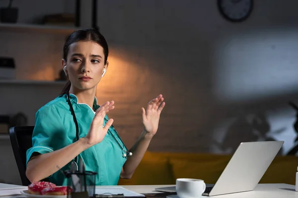 Attraktive Krankenschwester in Uniform, die mit Kopfhörern am Tisch sitzt und während der Nachtschicht wegschaut — Stockfoto